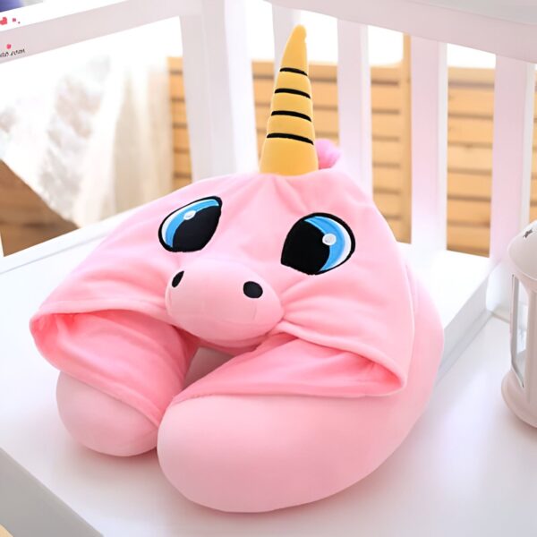 Image d'un coussin de voyage en forme de licorne rose avec des yeux bleus, un nez noir et une corne jaune à rayures noires. Le coussin repose sur une surface blanche devant un fond de barrières blanches et un objet blanc sur le côté droit., 45501060841727 | Licorne Shop