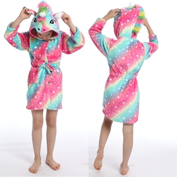 Une jeune fille porte une robe de chambre licorne rose et arc-en-ciel avec une capuche. La robe de chambre est à manches longues et a une ceinture à la taille. La capuche est doublée de polaire rose. La jeune fille est pieds nus et a les cheveux longs et blonds. Elle sourit et regarde l'appareil photo.
