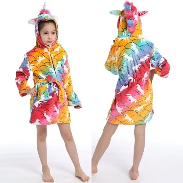 Une jeune fille porte une robe de chambre licorne arc-en-ciel avec une capuche. La robe de chambre est jaune avec des écailles arc-en-ciel et une doublure en polaire arc-en-ciel. La capuche est dotée d'une corne de licorne et d'oreilles de licorne. La jeune fille a les cheveux longs et blonds et elle sourit.