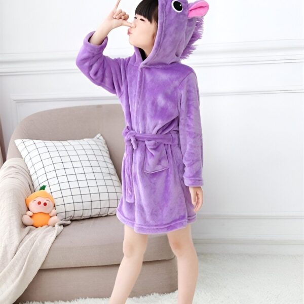 Une jeune fille porte une combinaison pyjama licorne violette avec une capuche et une ceinture. Elle se tient debout sur un tapis beige dans un salon.