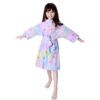 peignoir-licorne-fille-etoile-5a.jpg Une jeune fille porte une robe de chambre licorne violette avec des étoiles multicolores. Elle a les cheveux bruns et les yeux marrons. Elle est pieds nus et se tient debout, les bras ouverts.