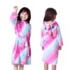 peignoir-licorne-fille-galaxie-3a.jpg Une petite fille porte une robe de chambre licorne rose et bleue à capuche. La robe de chambre est ouverte, on peut voir qu'elle porte un pyjama licorne rose et blanc. Elle a les cheveux bruns et les yeux marron. Elle est souriante et joyeuse.