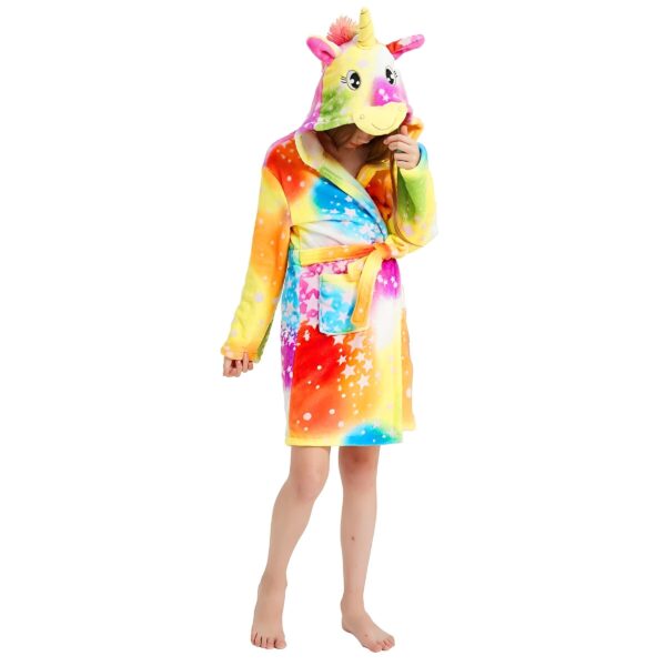 Une jeune fille porte une robe de chambre licorne arc-en-ciel avec une capuche. La robe de chambre est en peluche et a des étoiles blanches. La capuche a une corne de licorne et des oreilles. La jeune fille est pieds nus et a les cheveux longs et blonds.