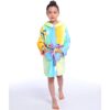 Une jeune fille porte une robe de chambre à capuche arc-en-ciel. La robe de chambre est jaune, rose, bleue et verte. La fille a les cheveux bruns et les yeux marron. Elle est pieds nus.
