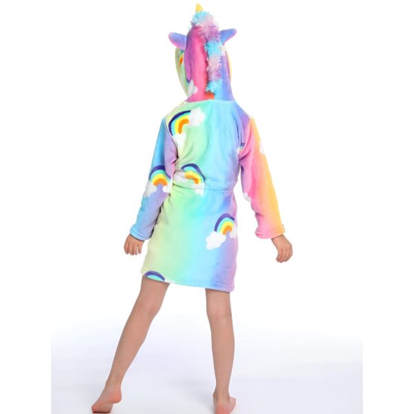 Une jeune fille porte une robe de chambre licorne arc-en-ciel avec une capuche. La robe de chambre est rose, violette et bleue avec des nuages blancs et des arcs-en-ciel. La capuche est en forme de tête de licorne avec une corne et des oreilles.