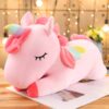 Une peluche licorne rose allongée sur une table en bois. La peluche a une crinière et une queue arc-en-ciel, ainsi que des ailes multicolores.