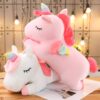 Une peluche licorne rose et blanche allongée sur le sol. La peluche licorne rose a une crinière et une queue arc-en-ciel. La peluche licorne blanche a une crinière et une queue roses. Les deux peluches ont les yeux fermés.