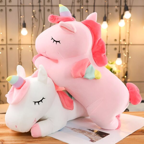Une peluche licorne rose et blanche allongée sur le sol. La peluche licorne rose a une crinière et une queue arc-en-ciel. La peluche licorne blanche a une crinière et une queue roses. Les deux peluches ont les yeux fermés.