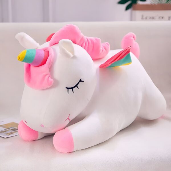 peluche-licorne-couchee-blanche-1a-1.jpg Une peluche licorne blanche et rose allongée sur un lit avec les yeux fermés. La peluche a une crinière et une queue roses, ainsi qu'une corne multicolore.