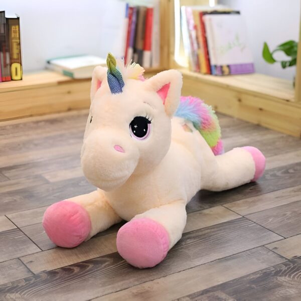 Une peluche licorne allongée sur le sol. La peluche est blanche avec une crinière et une queue arc-en-ciel. Elle a des yeux roses et des sabots roses.