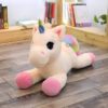 Une peluche licorne allongée sur le sol. La peluche est blanche avec une crinière et une queue arc-en-ciel. Elle a des yeux roses et des sabots roses.