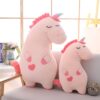 Deux peluches licornes roses, une grande et une petite, sont assises sur un canapé gris clair devant une fenêtre ensoleillée. Les licornes ont des crinières roses, des cornes multicolores et des cœurs roses sur leur corps., 45501421551871 | Licorne Shop