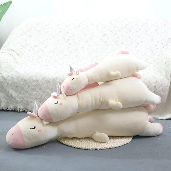 Une image de trois peluches de licorne blanche et rose allongées sur un tapis et un canapé. Les peluches ont des cornes argentées et des ailes roses.