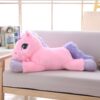 peluche-licorne-rose-1a.jpg Une peluche géante de couleur rose et violet en forme de licorne allongée sur un canapé beige dans un salon moderne.