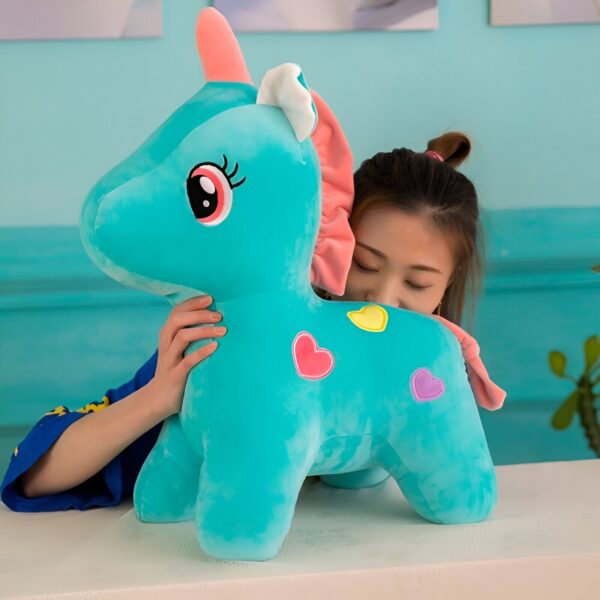 peluche-licorne-verte-1a.jpg Une peluche licorne bleue avec une crinière et une queue arc-en-ciel. La peluche est assise sur une surface blanche et est entourée de coeurs en feutrine de couleur rose, jaune et bleu ciel.