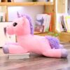 peluche-licorne-xxl-rose-1a.jpg Une peluche licorne rose et violette allongée sur le sol devant une bibliothèque.