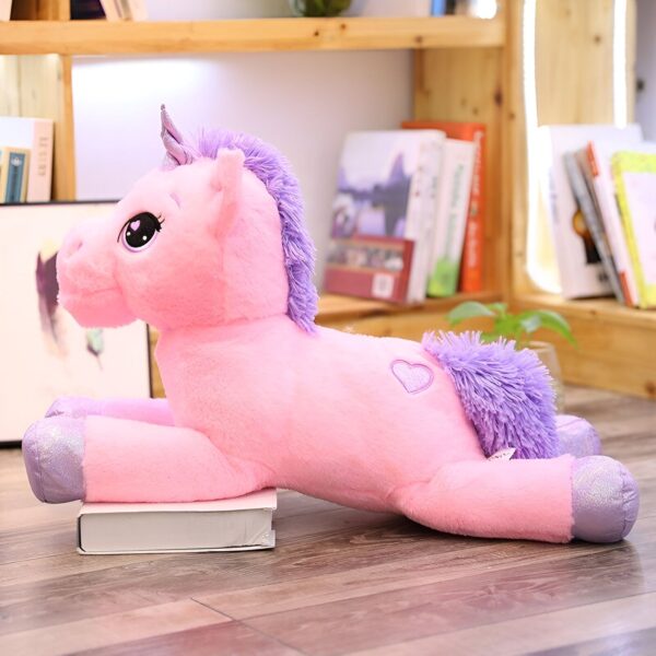 peluche-licorne-xxl-rose-1a.jpg Une peluche licorne rose et violette allongée sur le sol devant une bibliothèque.