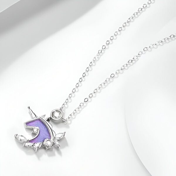 Un collier avec un pendentif en forme de licorne. La licorne est violette avec une crinière et une queue roses. Le pendentif est entouré de feuilles et de fleurs. La chaîne est en argent.