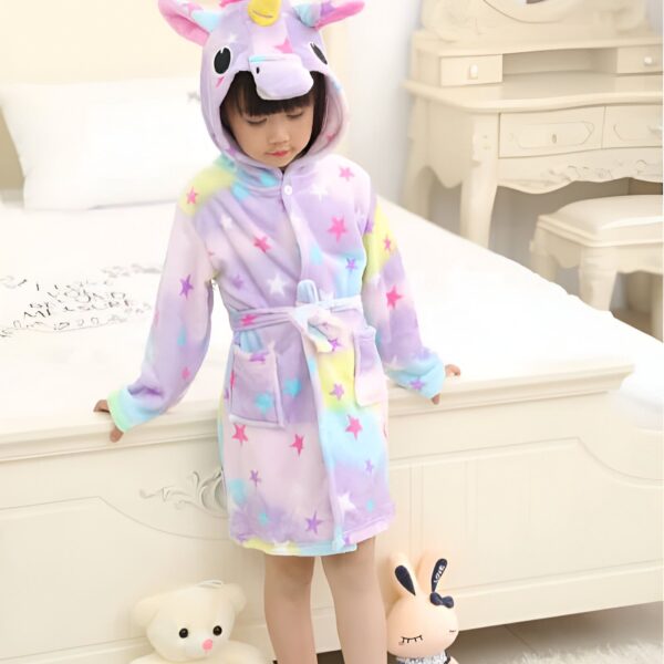 Une petite fille porte un peignoir licorne coloré avec des étoiles, debout près d'un lit avec des jouets., 45502344986879,45502345019647,45502345052415,45502345085183,45502345117951,45502345150719,45502345183487,45502345216255 | Licorne Shop