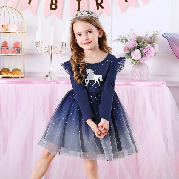 Une petite fille porte une robe bleue à manches longues avec une licorne imprimée sur le devant. La robe est ornée de tulle pailleté argenté et d'un nœud rose à la taille. La petite fille est coiffée d'une couronne argentée et porte des chaussures roses. Elle se tient debout devant un mur rose et blanc décoré de ballons et de banderoles.