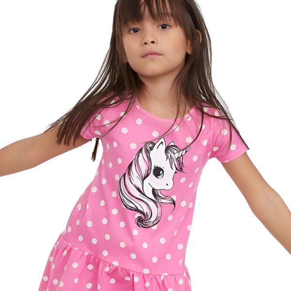 Une petite fille porte une robe rose à pois blancs avec une image de licorne. La licorne est blanche avec une crinière et une queue roses.