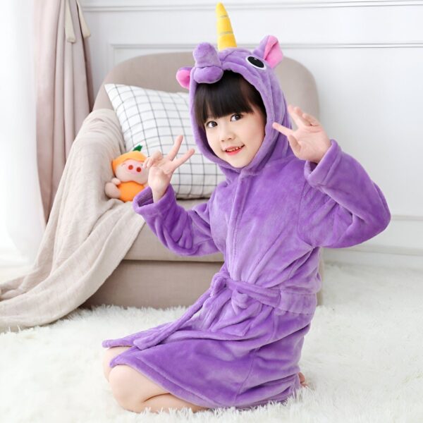 Une petite fille souriante à genoux sur un tapis blanc, portant un peignoir violet à capuche avec une corne de licorne et des oreilles roses. Elle fait un signe de paix avec ses mains. Derrière elle, on aperçoit un canapé, un coussin et une peluche orange., 45502350196991,45502350229759,45502350262527,45502350295295,45502350328063,45502350360831,45502350393599,45502350426367 | Licorne Shop