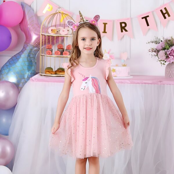 robe-licorne-rose-fille-courte-1a.jpg Une petite fille porte une robe rose à motifs de licorne. Elle a un serre-tête avec des oreilles de licorne et des ailes de papillon. Elle est entourée de ballons et de décorations de fête.