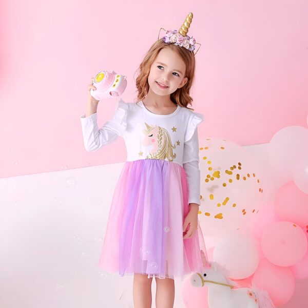 Une petite fille porte une robe rose et blanche à manches longues avec une licorne imprimée sur le devant. Elle porte également un serre-tête avec une corne de licorne et des fleurs. Elle tient un appareil photo rose à la main et sourit.