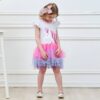Une petite fille porte une robe blanche à manches courtes avec une licorne imprimée sur le devant. La robe est également dotée d'un jupon en tulle rose, violet et bleu.