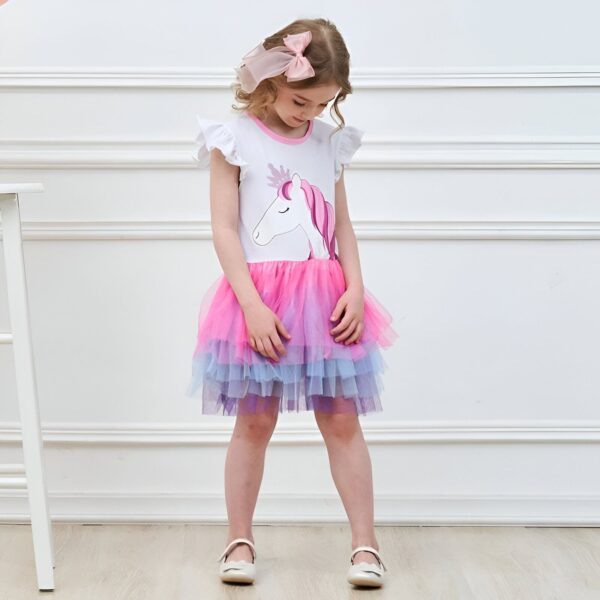Une petite fille porte une robe blanche à manches courtes avec une licorne imprimée sur le devant. La robe est également dotée d'un jupon en tulle rose, violet et bleu.