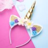 Serre-tête licorne avec corne dorée, oreilles blanches à paillettes dorées et fleurs blanches, bleues et violettes. Fond rose et bleu pastel., 45501336453375 | Licorne Shop