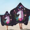 serviette-de-plage-motif-licorne-australienne-1a.jpg Deux personnes sur une plage portant des serviettes à capuche à motif de licorne. Les serviettes sont noires et décorées de licornes blanches avec des crinières roses et de fleurs colorées. Le texte "unicorns are real" est visible., 45501225238783 | Licorne Shop