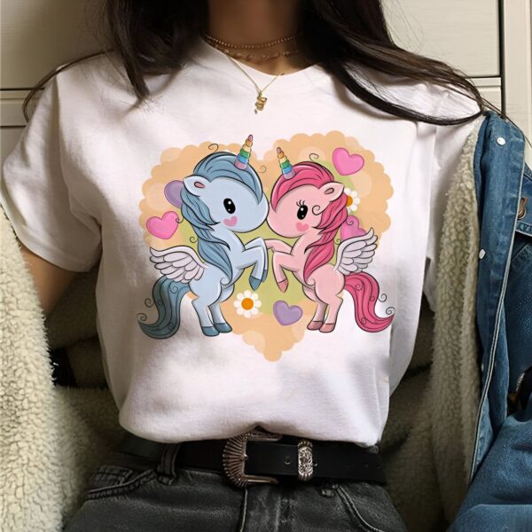 t-shirt-licorne-couple-amoureux-1a.jpg Une jeune femme porte un t-shirt blanc avec un motif de deux licornes. Les licornes sont de couleur rose et bleue et ont des ailes. Elles se tiennent debout sur leurs pattes arrières et se tiennent les mains. Il y a un cœur jaune en arrière-plan.