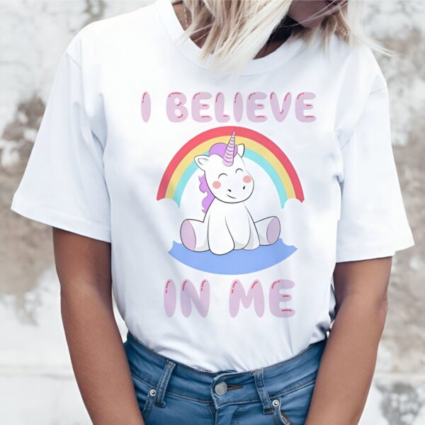 Une jeune femme porte un t-shirt blanc avec un arc-en-ciel et une licorne. La licorne est assise sur un nuage et le texte "I believe in me" est écrit en dessous.
