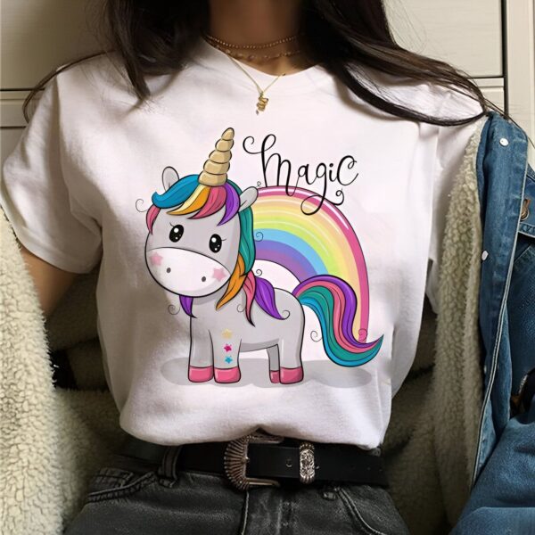 Une jeune femme porte un t-shirt blanc avec une image de licorne. La licorne est blanche avec une crinière et une queue arc-en-ciel. Il y a aussi un arc-en-ciel au-dessus de la licorne. Le texte "Magic" est écrit en noir au-dessus de la licorne.