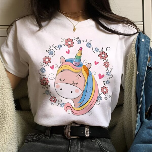 Une jeune femme porte un t-shirt blanc avec un motif de licorne. La licorne a une crinière et une queue arc-en-ciel, et est entourée de fleurs et de cœurs.