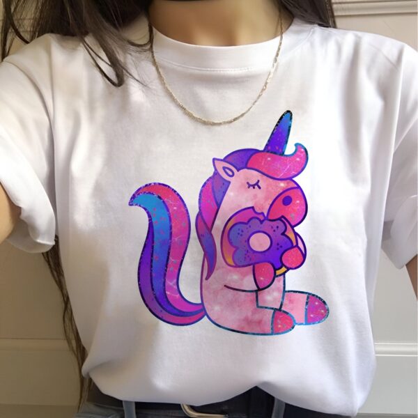 t-shirt-licorne-gourmande-1a.jpg Une illustration d'une licorne rose et violette tenant un beignet dans sa bouche. La licorne est assise sur un fond blanc.