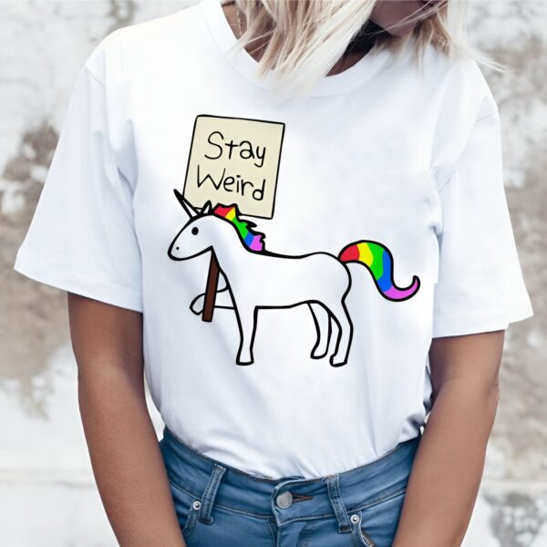 Une femme porte un t-shirt blanc avec un dessin d'une licorne tenant une pancarte avec le texte "Stay Weird". La licorne a une crinière et une queue arc-en-ciel.