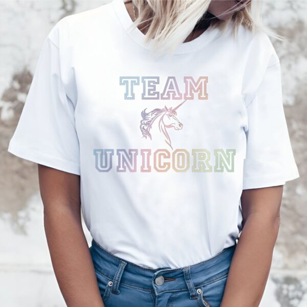 Une jeune femme porte un t-shirt blanc avec le texte "Team Unicorn" écrit dessus. Le texte est coloré avec un dégradé de couleurs arc-en-ciel. La femme est blonde et porte un jean bleu. Elle est souriante et regarde l'appareil photo.