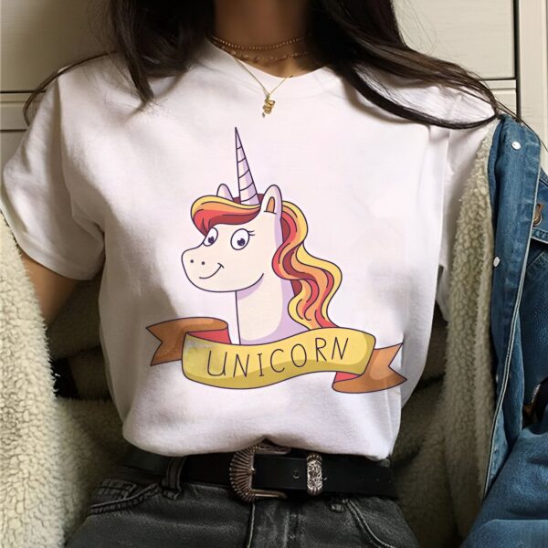 t-shirt-licorne-unicorn-1a_6250cd1a-03ab-4117-a2cf-cf053e1cc95d.jpg Une jeune femme porte un t-shirt blanc avec un dessin de licorne. La licorne a une crinière et une queue arc-en-ciel. Le texte "UNICORN" est écrit en lettres jaunes sur une banderole rose.