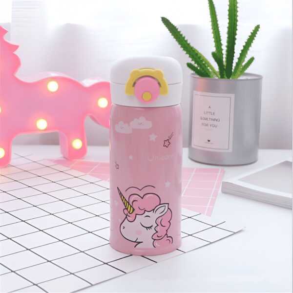 tasse-de-voyage-licorne-isotherme-pour-enfant-1a.jpg Gourde rose pour enfants avec motif de licorne et bouchon blanc, posée sur une table à carreaux. À l'arrière-plan, on aperçoit une lampe en forme de licorne illuminée et une plante verte dans un pot., 45501193257215 | Licorne Shop
