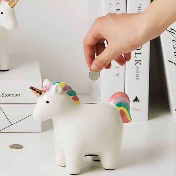 tirelire-licorne-enfant-en-ceramique-3a Une tirelire en céramique blanche en forme de licorne. La licorne a une corne dorée, une crinière et une queue arc-en-ciel. La tirelire est posée sur une étagère blanche.