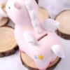 tirelire-licorne-kawaii-rose-en-resine-2a Une tirelire en forme de licorne rose avec des ailes blanches et une crinière blanche. La licorne est debout sur une bûche de bois.
