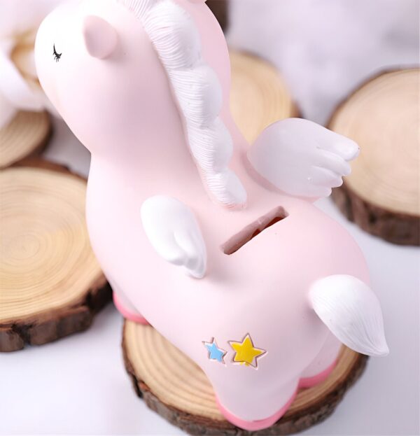 tirelire-licorne-kawaii-rose-en-resine-2a Une tirelire en forme de licorne rose avec des ailes blanches et une crinière blanche. La licorne est debout sur une bûche de bois.