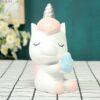 tirelire-licorne-mignonne-rose-en-resine-1a Une figurine de licorne blanche et rose tenant un oreiller bleu avec le mot "love" dessus. La licorne a les yeux fermés et un sourire sur le visage. Elle est assise sur une table à carreaux noirs et blancs. En arrière-plan, on peut apercevoir un mur vert et des fleurs blanches.