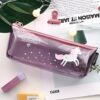 trousse-licorne-transparente-violette-1a.jpg Une trousse transparente avec une licorne imprimée dessus. La trousse est remplie de fournitures scolaires, dont des crayons, des stylos et une gomme.