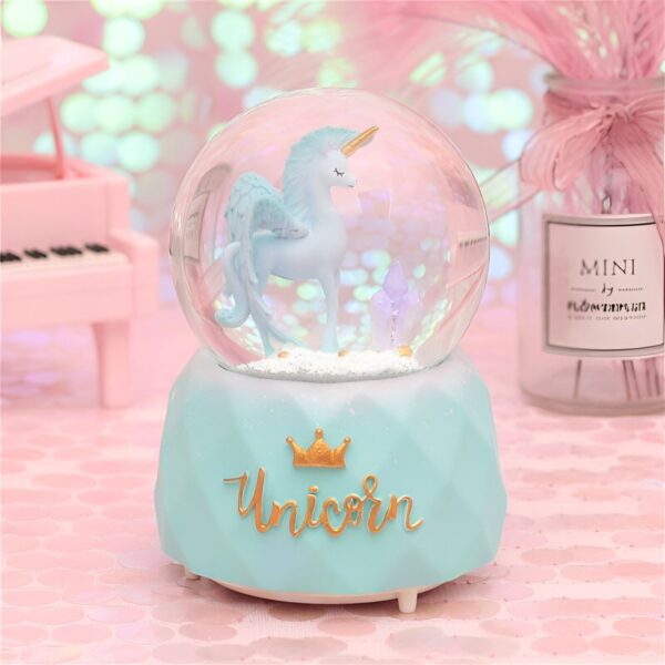 une-boule-a-neige-magique-licorne-bleue-1a.jpg Boule à neige féérique avec une figurine de licorne ailée à l'intérieur, posée sur un socle turquoise orné d'une couronne dorée et du mot "Unicorn" en lettres dorées. Un piano miniature rose et un vase décorent l'arrière-plan., 45501133521151 | Licorne Shop